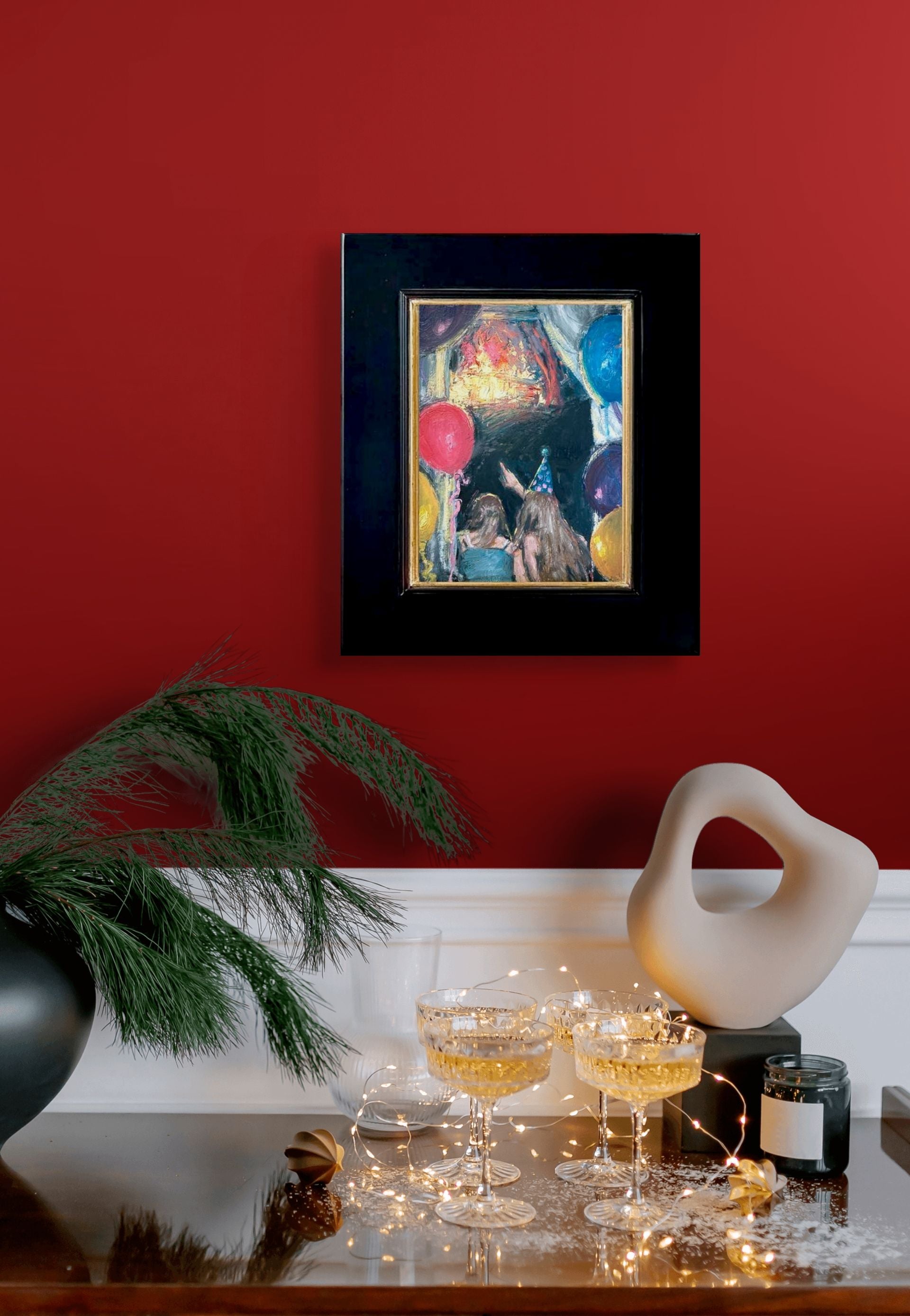 Impressionistic depiction of two people with party hats and balloons against a dark background looking and pointing at a lighted stage; shown with 2" black wood frame and gold lining; shown on red wall above shelf with tiny lights and bubbly drinks