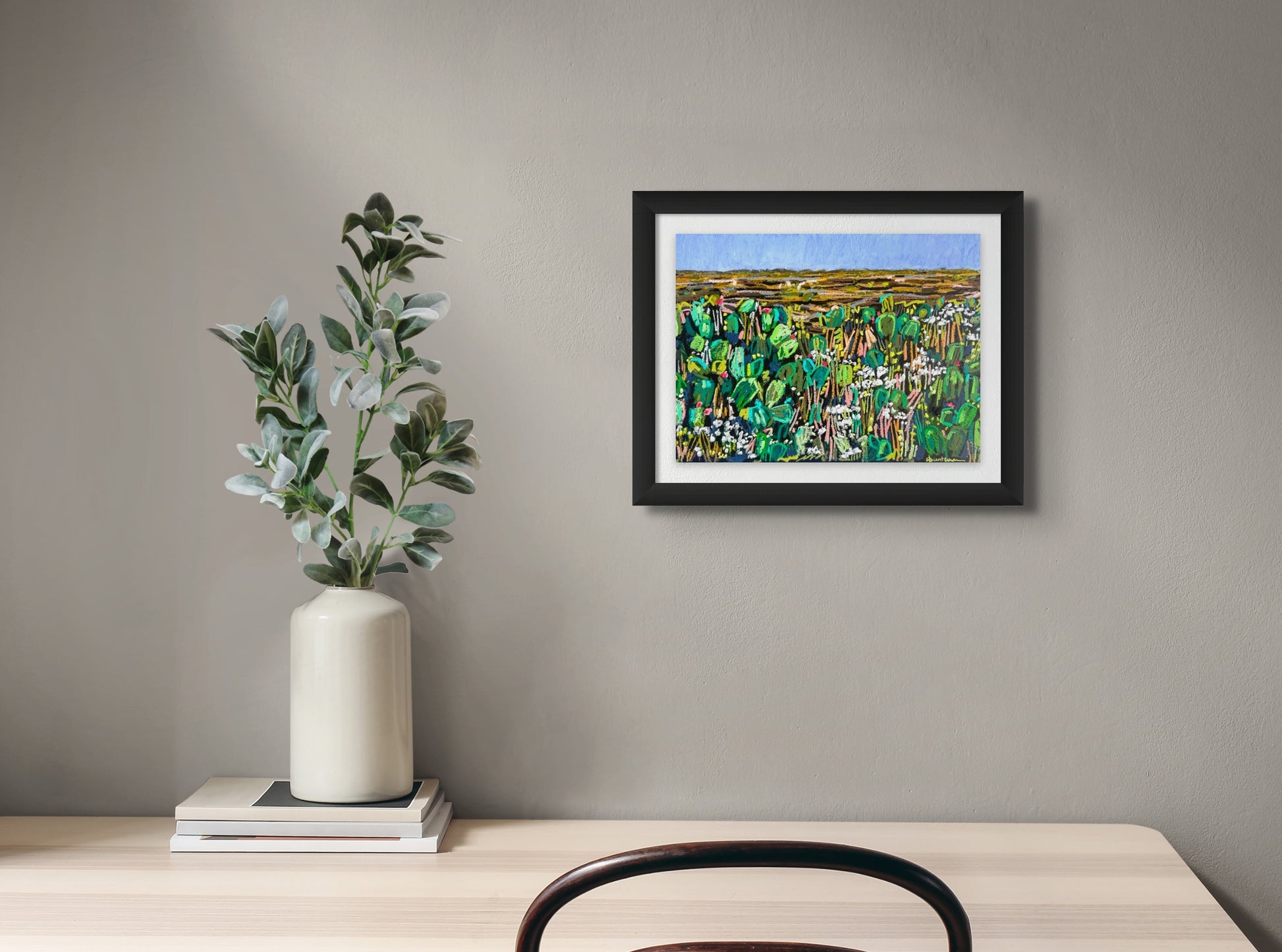 Oil pastel painting shows a cactus garden in the forefront in hues of greens and browns. In the distance is a brown landscape; artist Rachel Bunteman; shown in situ with white mat and black frame;