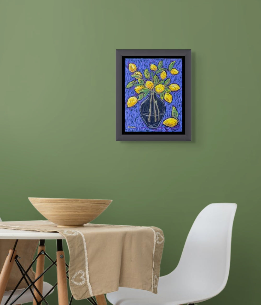 Bright and vivid oil pastel painting of clear vase filled with yellow lemons and their leaves against a blue background; artist Rachel Bunteman; 'Electric Lemons' hangs above a table and chair on a green wall. The painting is in a gray frame with a thin black mat.