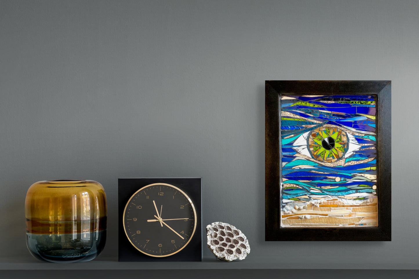 This stunning 11"x14" (with frame) mosaic piece is housed in a black frame. In the center is a green eye with touches of tans and lighter shades of green. The eye sits among a sea of blue and green glass that portray an ocean's current; shown on wall in situ; artist Denise Marshall