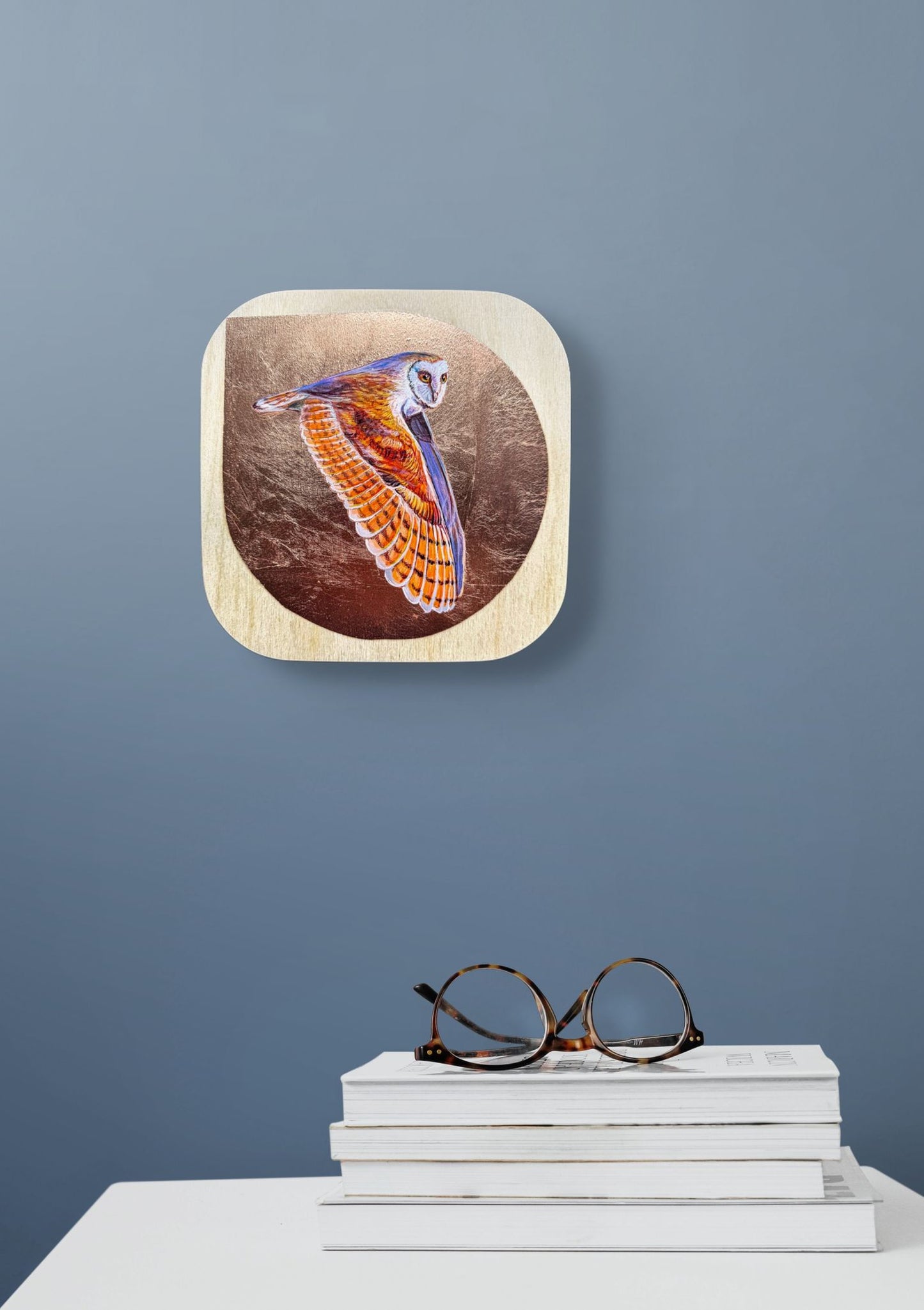 Colorful painted flying owl against copper leaf background on birch wood panel; 8"x8"; shown in situ on grey wall above white table, books, and pair of glasses; artist Marie Lavallee