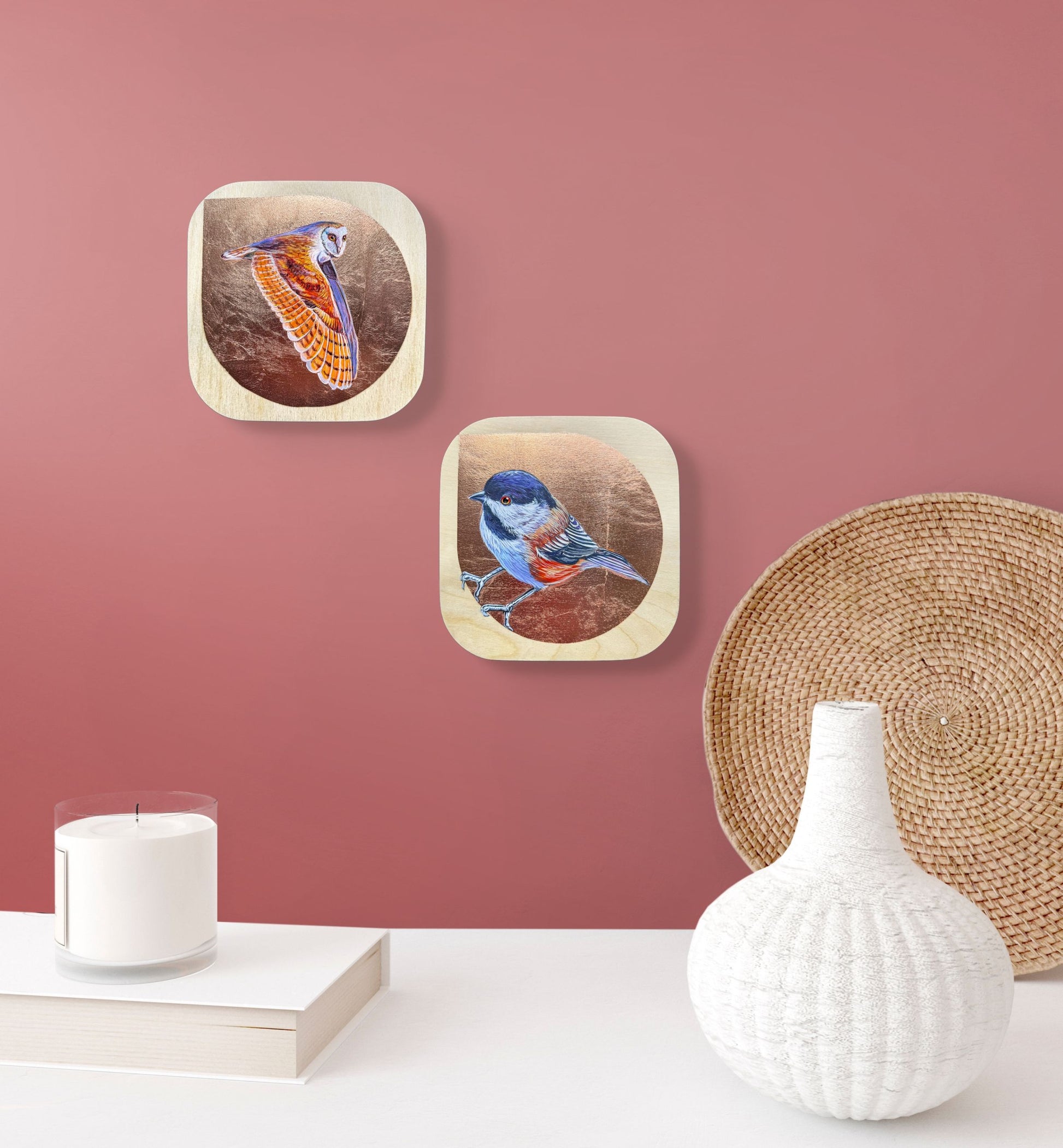 Two colorful paintings against copper leaf backgrounds on wood, one of a flying owl and one of a chickadee; hung as a pair on a rose-colored wall over a white table; artist Marie Lavallee