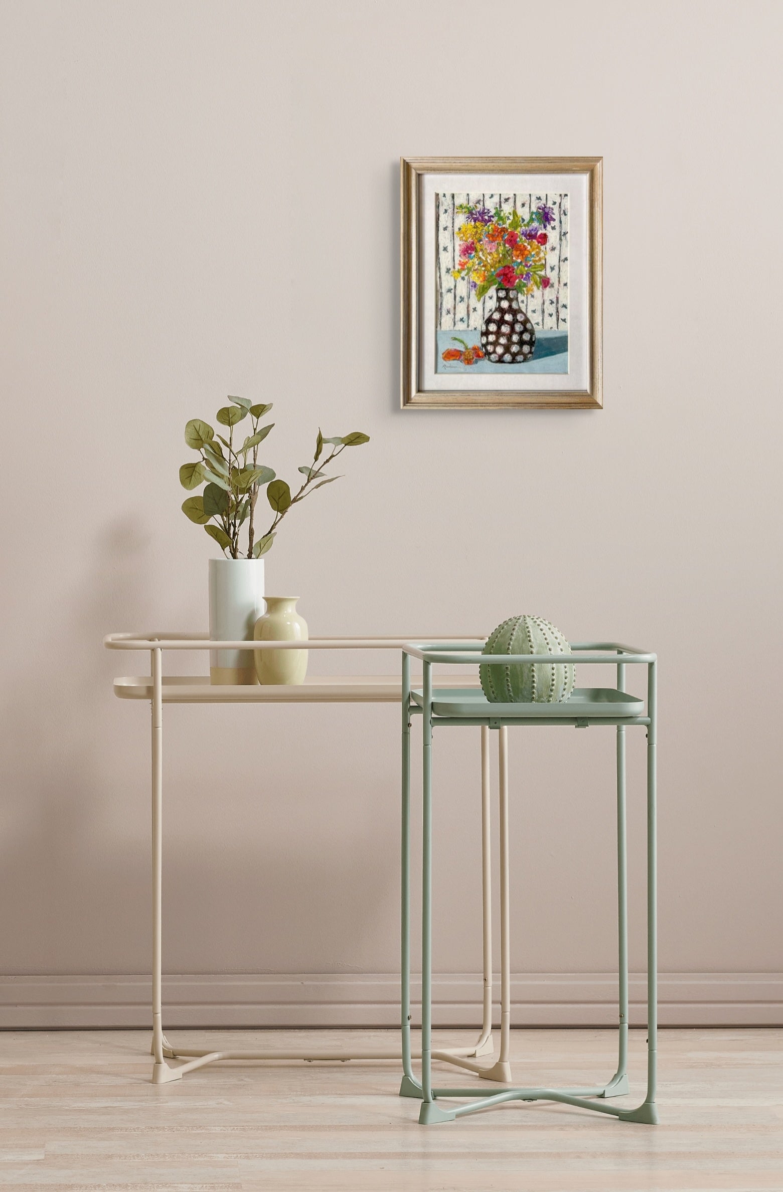 The painting hangs on a pale white wall over two small metal tables in ivory and mint green. A vase with leaves and two small empty vases sit on the tables. 