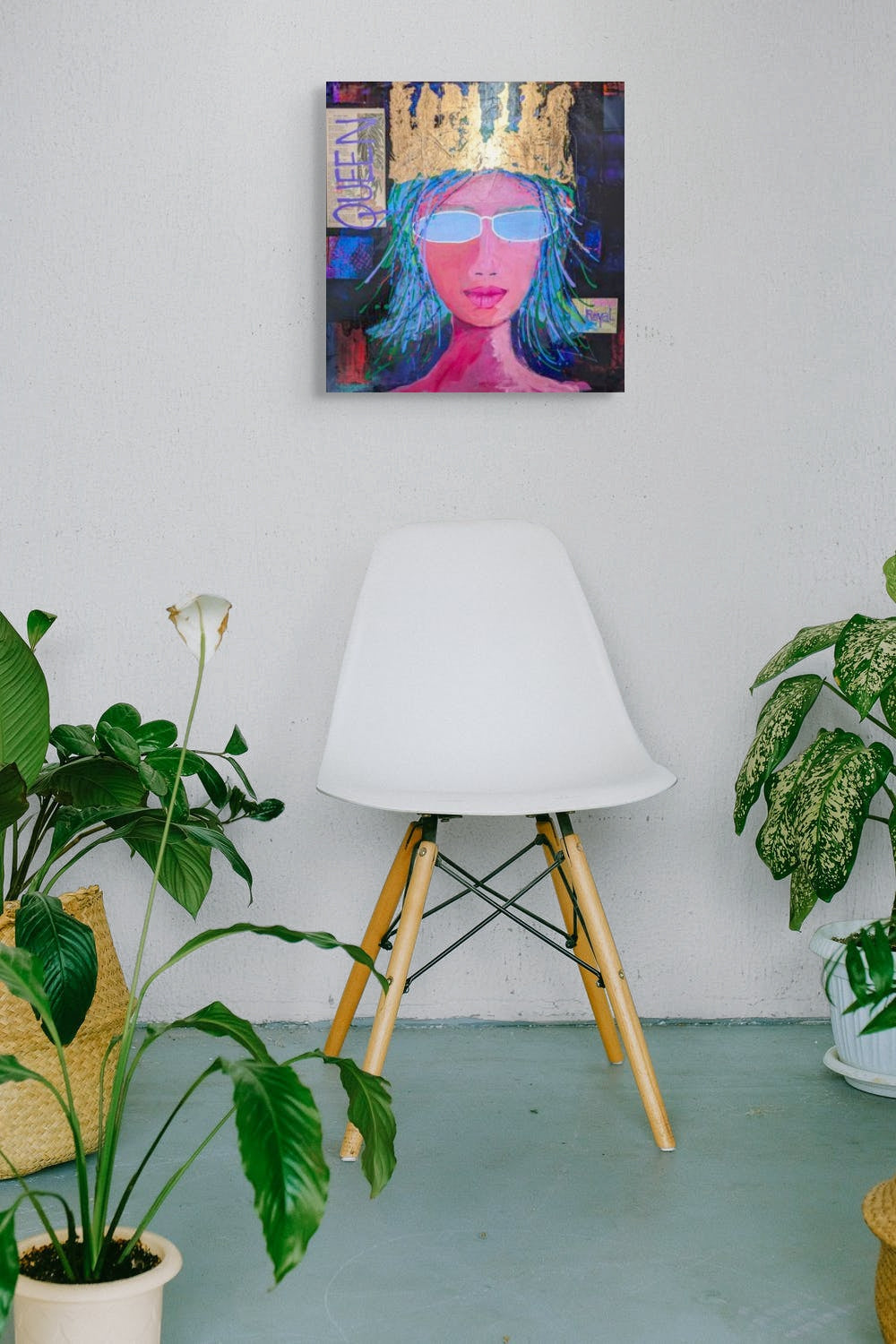 This in situ image of Her Majesty shows her reigning on a white wall above a modern design chair. Houseplants are on the floor near the chair. 