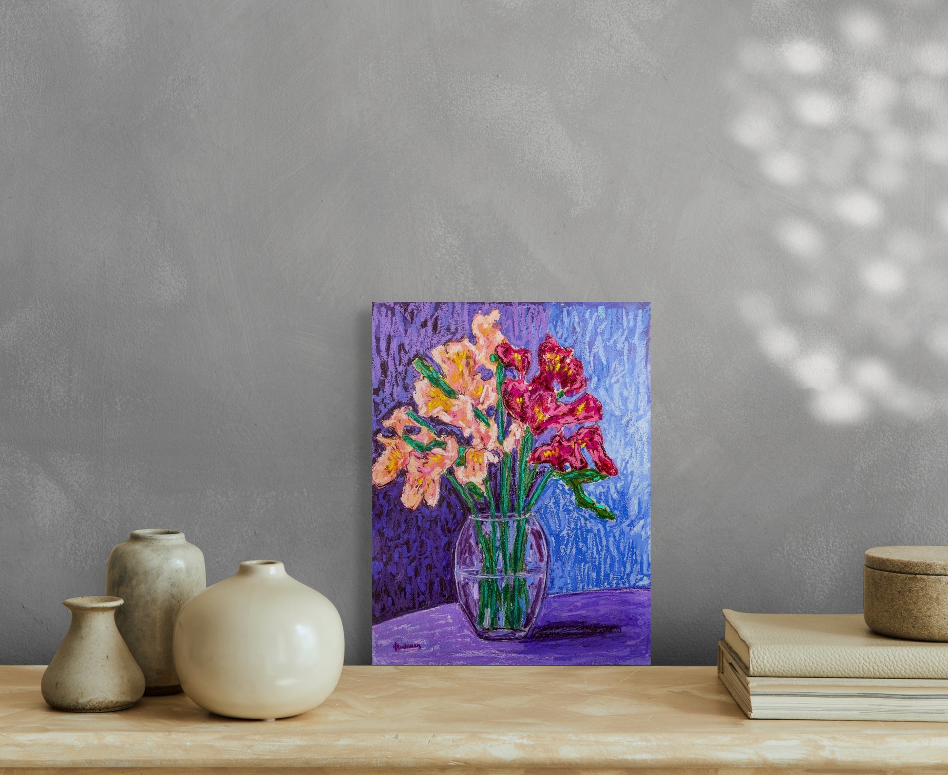 Bright and colorful oil pastel painting of clear glass vase filled with burgundy and peach Peruvian lilies against and blue and purple background; artist Rachel Bunteman; This in situ image shows the painting sitting on a table against a gray wall with sunshine and shadows. 