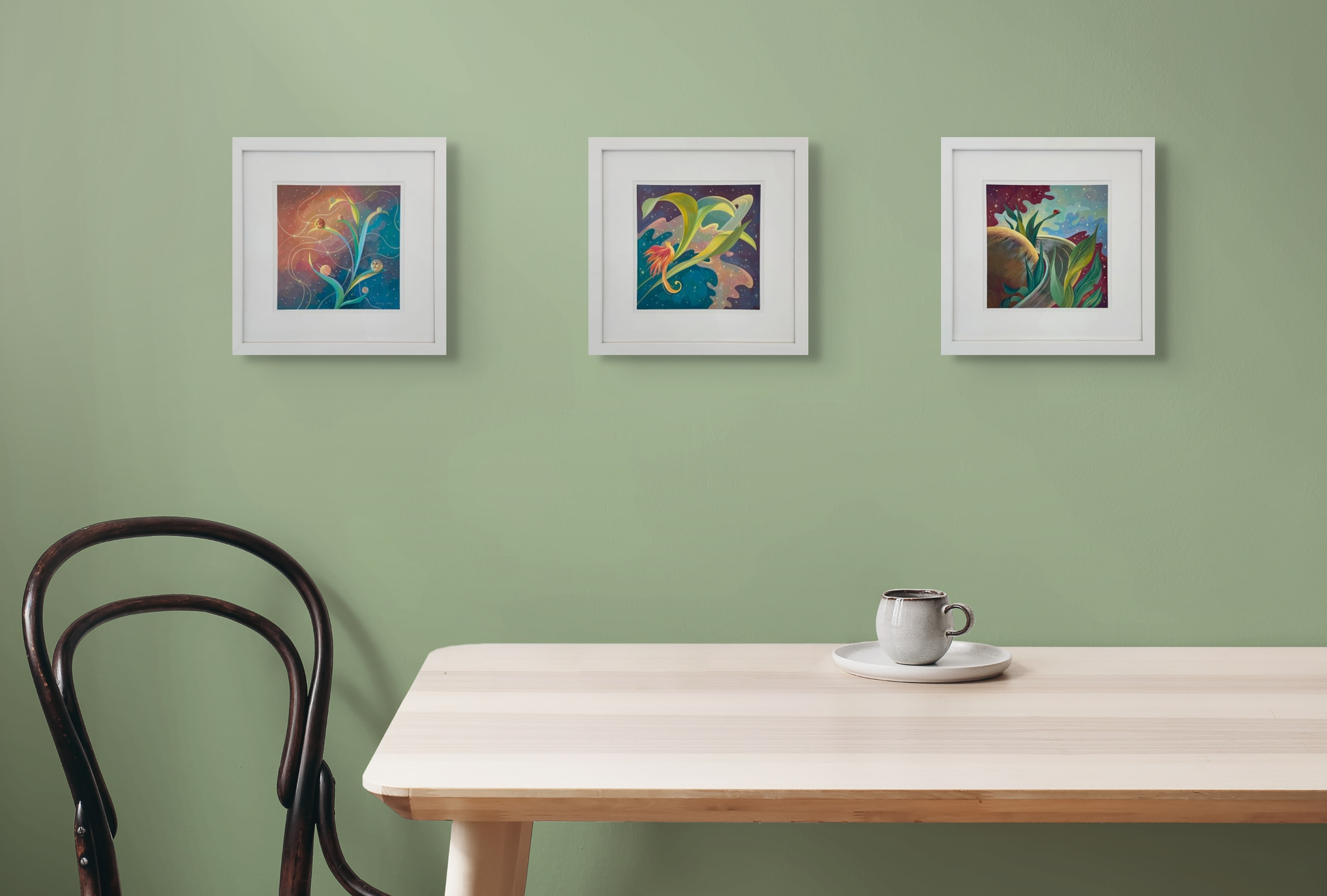 Three of Dany's framed pieces, Planetary Phases,  Saturn Feathers and Lady Rings  hang as a set on a light green wall above a light oak table. A mug sits on the table and a black chair is to the left. 
