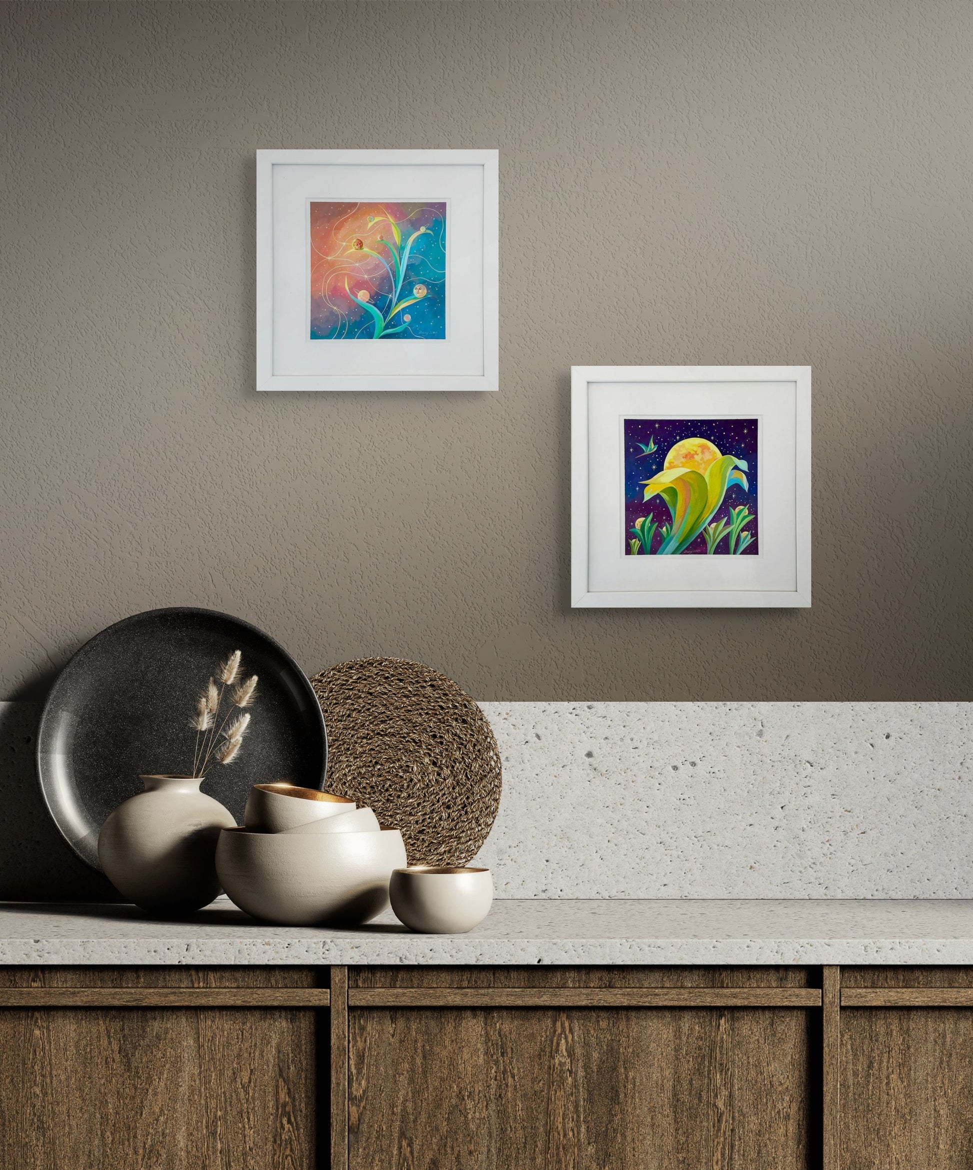 Two of Dany's pieces, Blooming Moons and Planetary Phases hang abouve a counter that is decorated with plates and bowls. The wall is a light gray and the cabinet brown wood with a white granite top. 