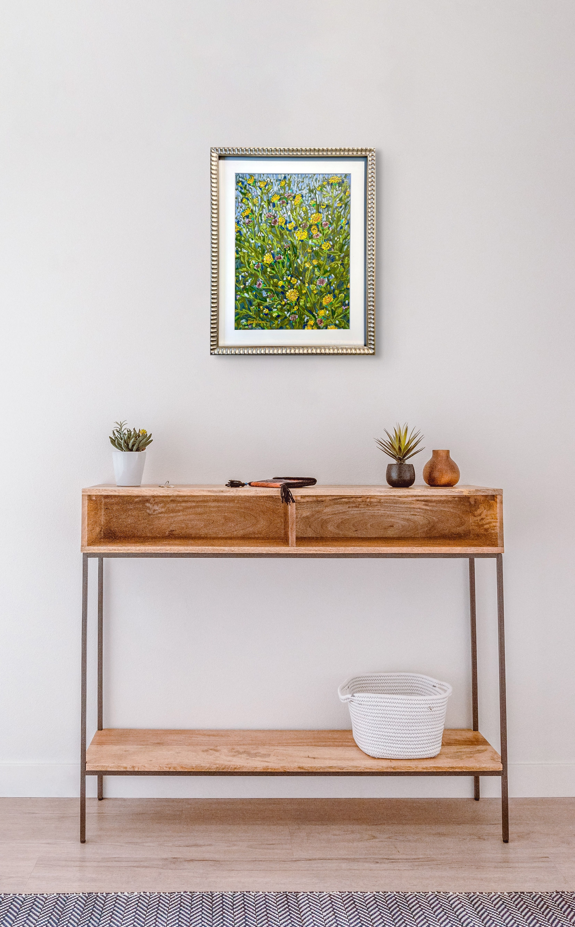 Queen For A Day hangs above a hallway table of light wood. Two plants sit on top of the table and blend well with the artwork