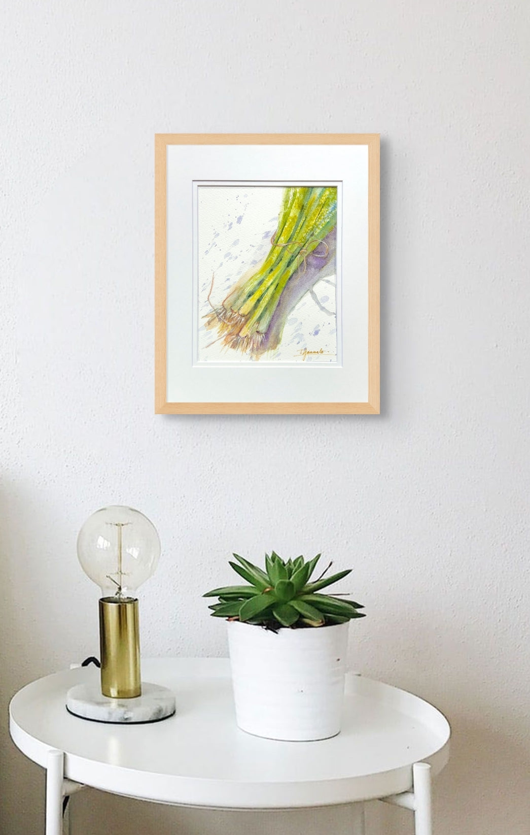 This still life of green onions brings a subtle touch of color to the wall. It hangs in a light colored wood frame over a small table with a lamp and a plant.