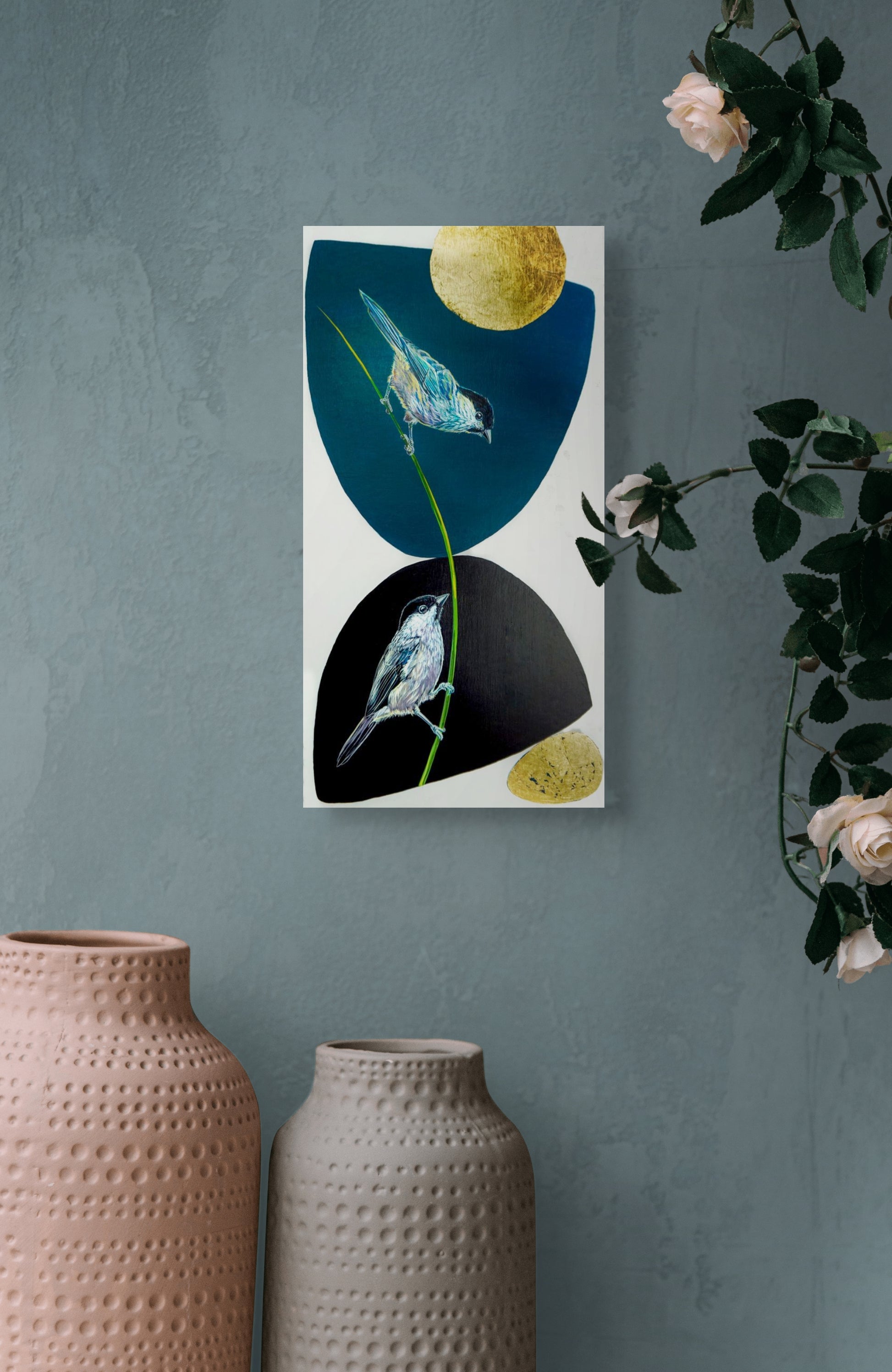 This in situ image shows Marie Lavallee's Balancing Act hanging on a blue gray wall over two large ceramic vases with roses on the edge of the photo. 